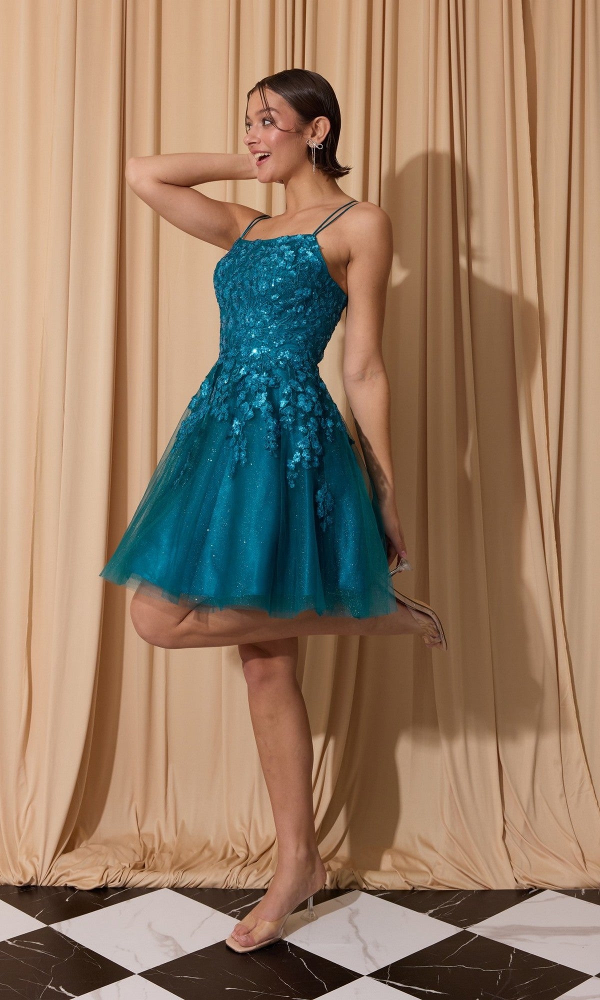 A woman smiles with one leg bent in front of beige curtains, wearing the Short Homecoming Dress: Eureka EK127 in teal, featuring a fit-and-flare silhouette with floral embellishments and a tulle skirt.